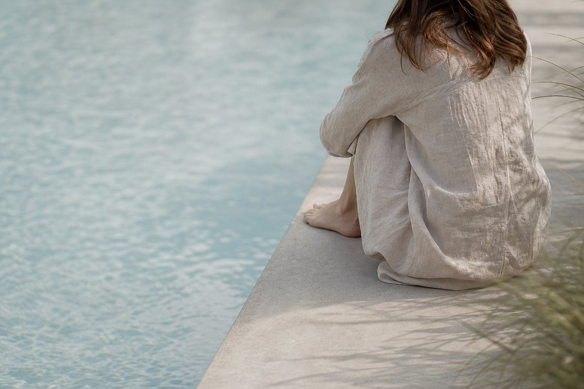 A woman in cream linen sits at a pool's edge with her knees drawn up and her back to the camera, dry grasses at the frame's edge.