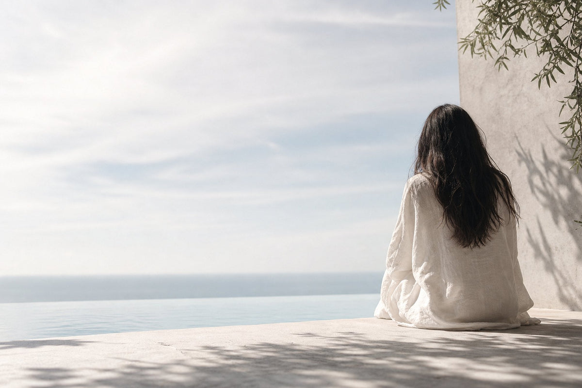 A woman with long dark hair sits on pale concrete facing the sea horizon, seen from behind, with olive foliage reaching into one corner of the frame.