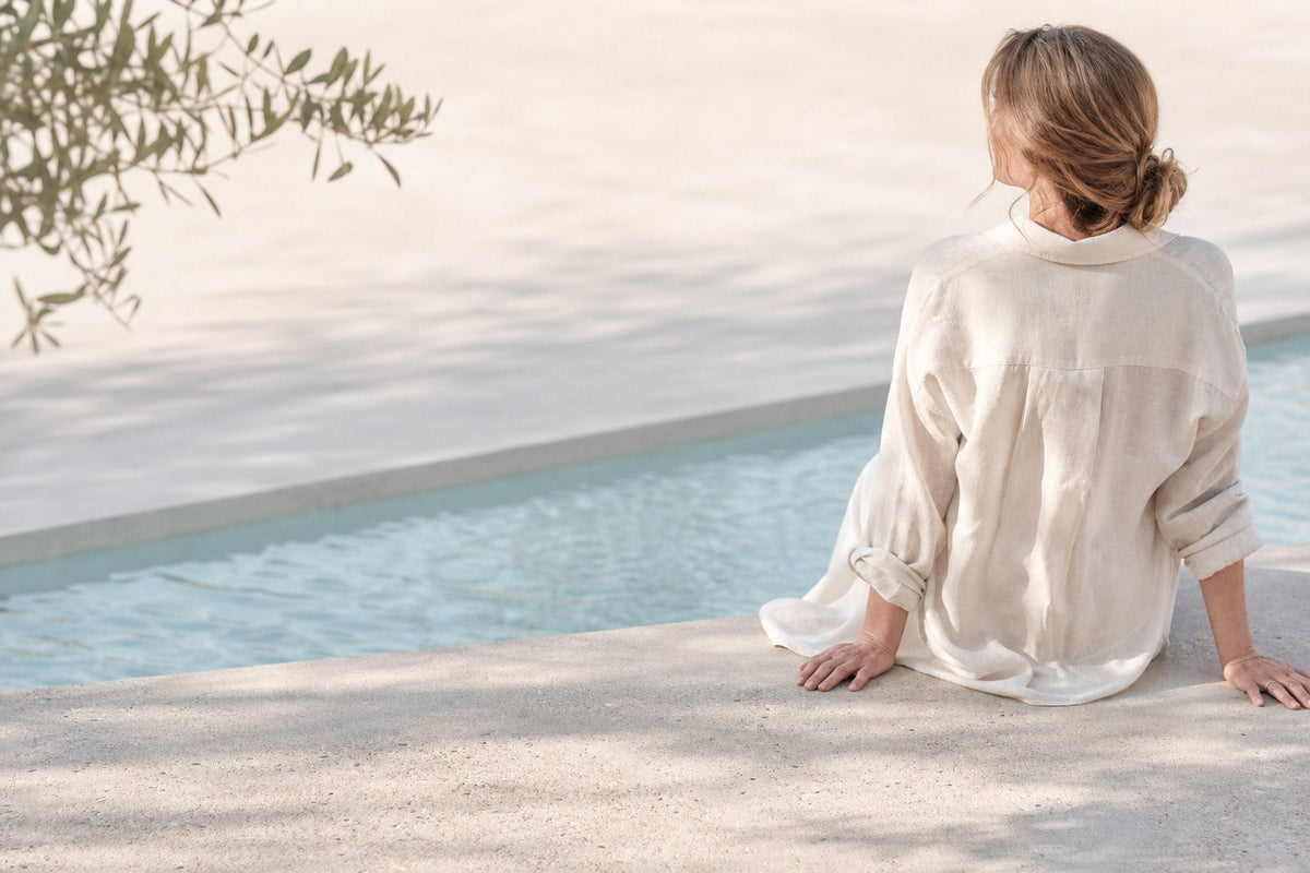 A woman sits at the edge of a pool seen from behind, in a loose cream linen shirt, with an olive branch overhead and sunlight falling on pale water.