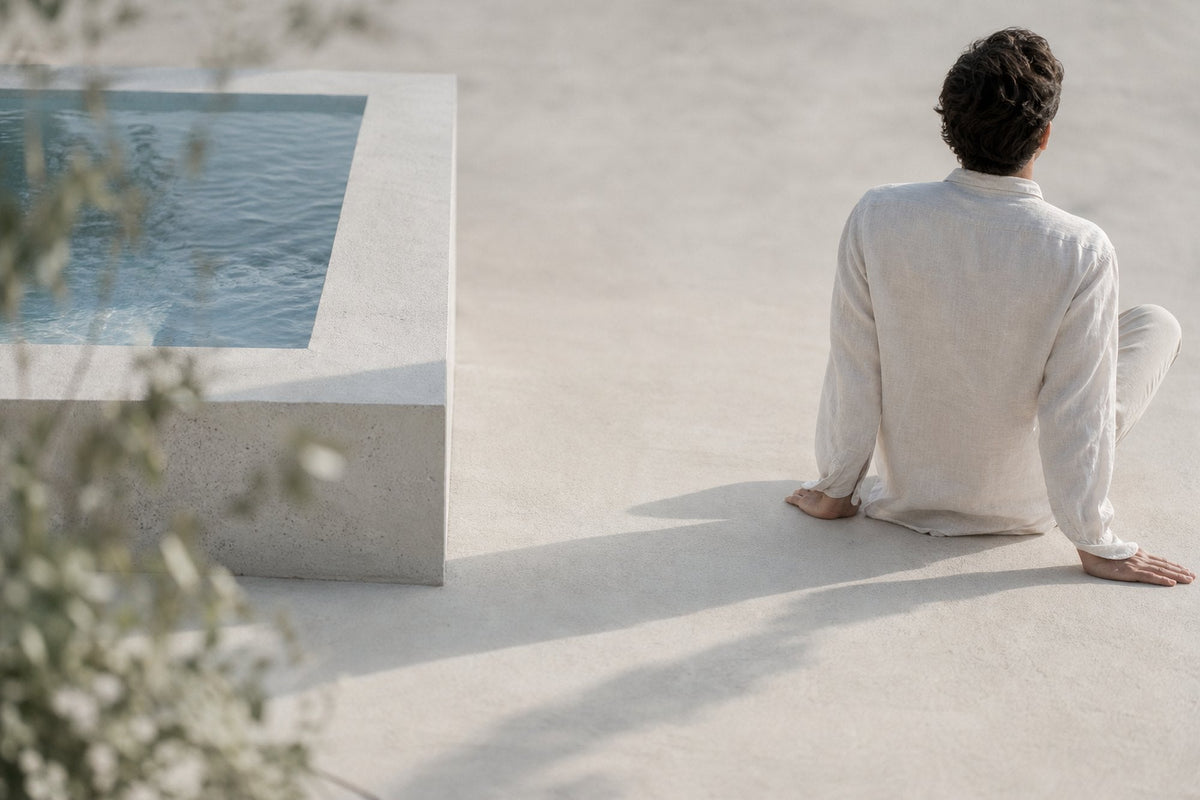 A man in a white linen shirt sits on broad concrete beside a raised pool, turned away from the camera, framed by soft olive foliage.