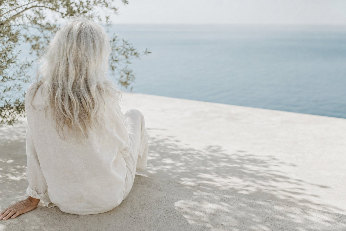 A woman with long silver hair sits on a pale terrace above the sea, seen from behind, in loose white linen with leaf shadows around her.