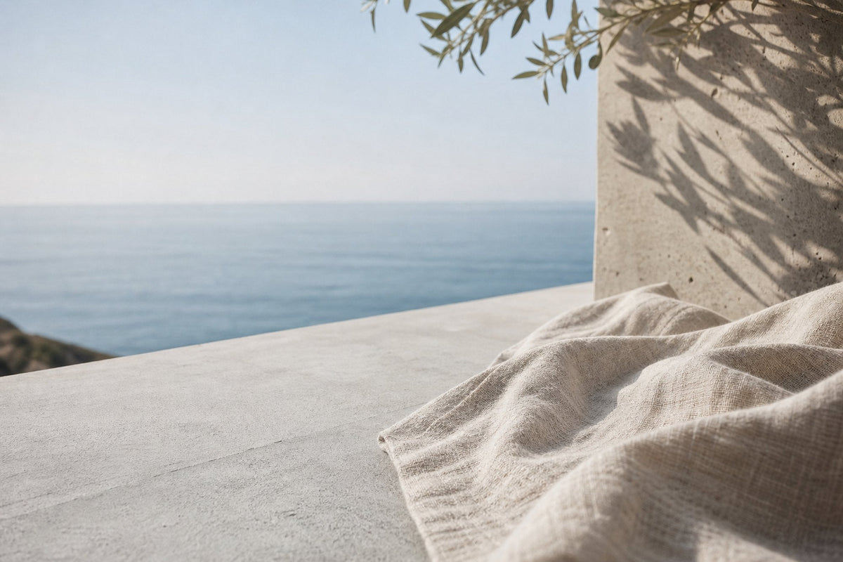 A loose linen throw lies folded on a pale concrete ledge above a calm sea, with olive-branch shadows falling across the wall behind it and no one in the frame.