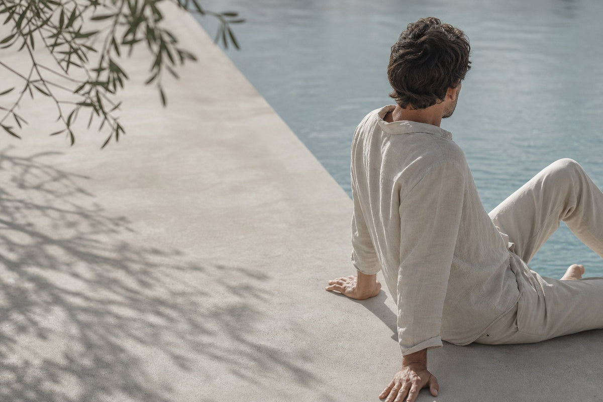 A man in loose linen sits on sunlit concrete beside still water, seen from behind, with olive foliage shading the left of the frame.