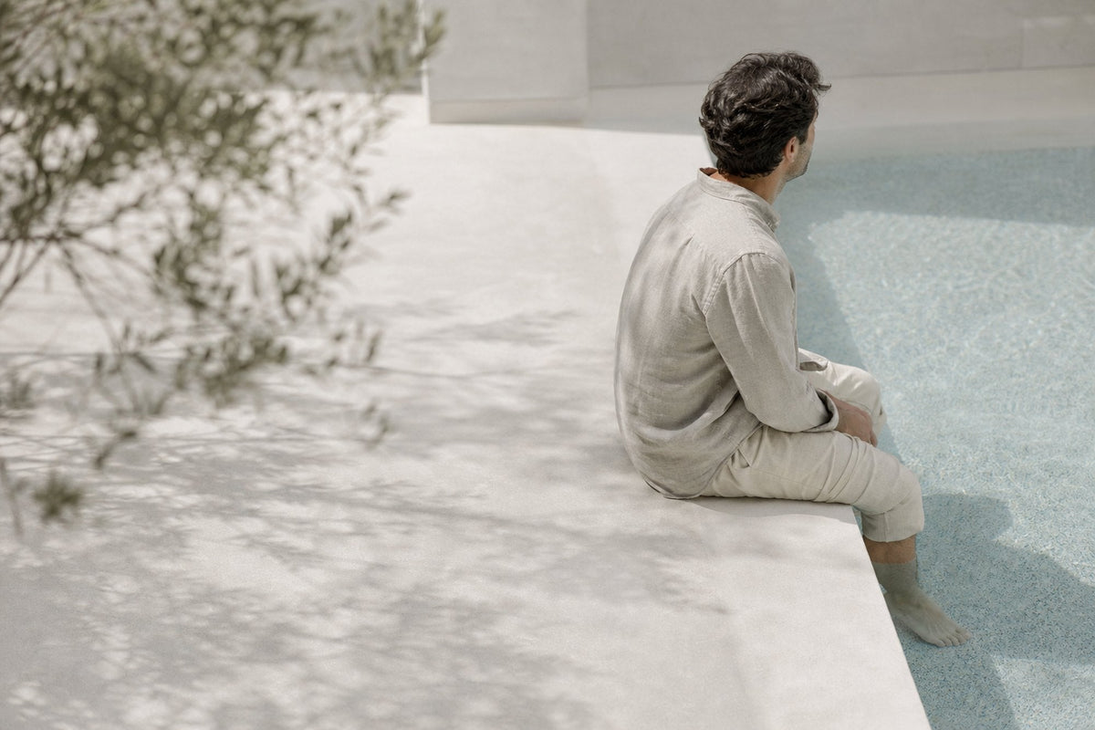 A man in cream linen sits at the edge of a pale pool with his back to the camera, olive branches and their shadows crossing the upper frame.