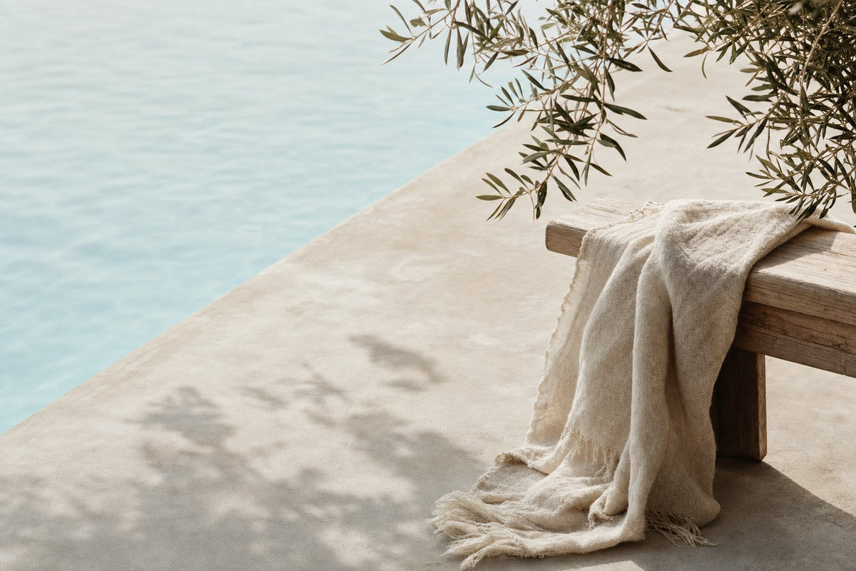 A linen throw draped over a wooden bench at the edge of a pale pool, with olive leaves and their shadows across the concrete.