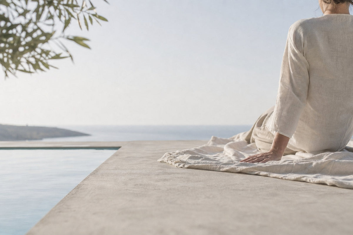 A woman sits on a linen throw at the edge of an infinity pool, seen from behind, looking out toward a pale sea and a distant headland.