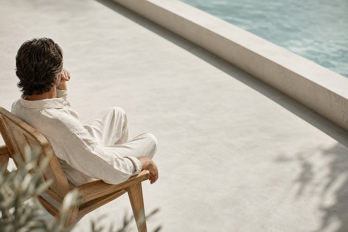 A man sits in a wooden chair beside a pool, seen from behind, in loose cream linen, with olive foliage in the foreground and sunlight across pale stone.