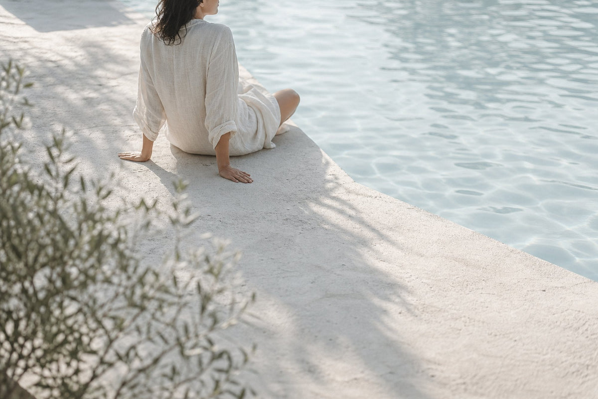 A woman in cream linen sits on a pale ledge at the edge of a pool, seen from behind, with olive-leaf shadows falling across the concrete.