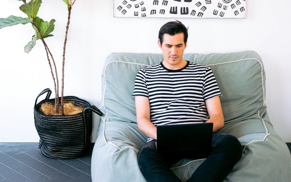 Man in striped shirt sitting on blue beanbag chair working on laptop
